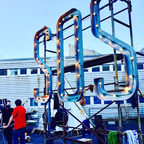 A large S.O.S. sign with light bulbs is mounted on a metal frame in front of a building with a corrugated metal exterior and windows. Two people stand nearby, one wearing a red shirt and the other with a face mask.