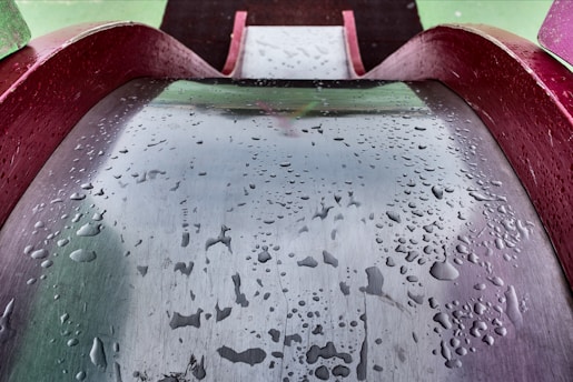 A metallic playground slide is wet with droplets of water scattered across its surface. The slide has a reflective quality, capturing hints of surrounding colors and shapes. It is bordered by maroon-colored edges and is set against a green background, implying an outdoor environment.