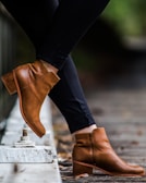 A person is wearing stylish brown leather ankle boots paired with black pants. The boots have a zip on the side and a low heel, standing out against a blurred background, likely an outdoor setting with a wooden path.