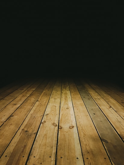 A perspective view of a wooden plank floor leading into a dark, shadowy background. The floor is composed of natural, textured wooden planks with visible grains and knots, creating a sense of depth and leading lines.