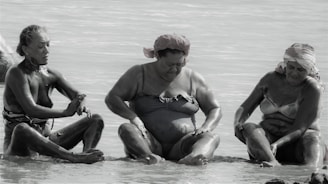 Three individuals sit by the sea, covered in mud, possibly engaging in a mud therapy or spa activity. They are in swimsuits and appear relaxed, with one person in the middle looking down, and the others on either side observing their hands or legs.