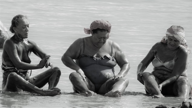 Three individuals sit by the sea, covered in mud, possibly engaging in a mud therapy or spa activity. They are in swimsuits and appear relaxed, with one person in the middle looking down, and the others on either side observing their hands or legs.