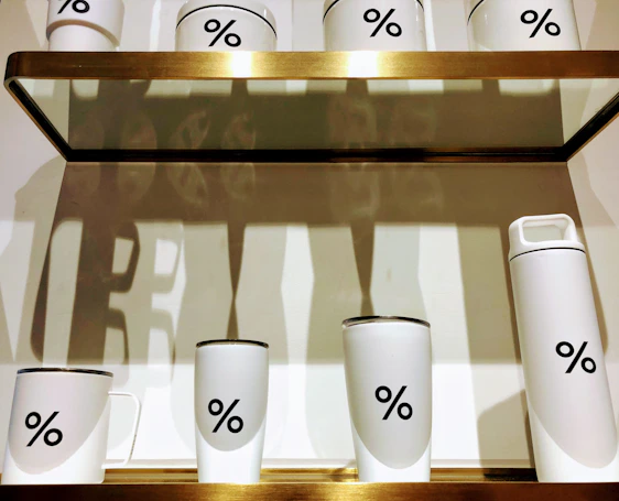 A collection of white cups and thermoses featuring the percentage symbol on a wooden shelf. The items create stark shadows against a white backdrop.