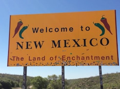 A large yellow sign with the text 'Welcome to New Mexico, The Land of Enchantment', featuring illustrations of red and green chili peppers. The sign is mounted on poles amidst a landscape of green vegetation and a clear blue sky.