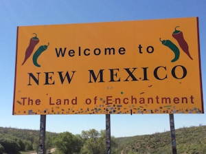 A large yellow sign with the text 'Welcome to New Mexico, The Land of Enchantment', featuring illustrations of red and green chili peppers. The sign is mounted on poles amidst a landscape of green vegetation and a clear blue sky.