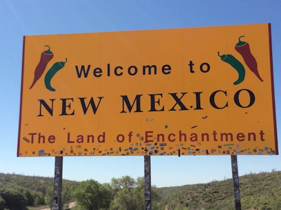 A large yellow sign with the text 'Welcome to New Mexico, The Land of Enchantment', featuring illustrations of red and green chili peppers. The sign is mounted on poles amidst a landscape of green vegetation and a clear blue sky.