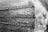 A black and white photograph showing a metal plaque or plate, possibly on a monument or directional indicator. It has engraved labels for various locations with associated numbers, which could represent altitudes or distances. The texture of the metal is rough and weathered, with some oxidization visible.