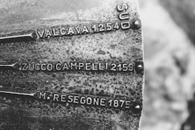 A black and white photograph showing a metal plaque or plate, possibly on a monument or directional indicator. It has engraved labels for various locations with associated numbers, which could represent altitudes or distances. The texture of the metal is rough and weathered, with some oxidization visible.