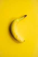A single banana positioned diagonally against a vibrant, solid yellow background. The banana has a natural curve and is ripe, featuring small brown spots near the stem.