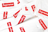 A collection of white plastic bags scattered around, each prominently featuring the red and white Supreme logo. The logo is centered on each bag, creating a repeated pattern throughout the image.