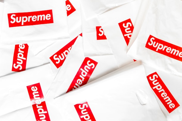 A collection of white plastic bags scattered around, each prominently featuring the red and white Supreme logo. The logo is centered on each bag, creating a repeated pattern throughout the image.
