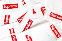 A collection of white plastic bags scattered around, each prominently featuring the red and white Supreme logo. The logo is centered on each bag, creating a repeated pattern throughout the image.