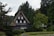 A traditional wooden house with a steep thatched roof is situated in a lush forested area. The house features multiple small windows and is surrounded by various green trees and shrubs. The background reveals tall evergreen trees, contributing to a serene and natural environment.