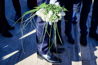 A person in formal attire holds a bouquet of white flowers and green foliage. The surrounding area features several other people dressed in dark clothing, with shadows cast on the ground.