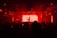 A dimly lit club scene with a DJ silhouetted against a bright red backdrop. The room is filled with people enjoying the atmosphere, and various speakers and lights are visible.
