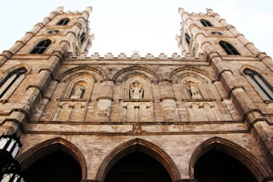 A majestic Gothic-style stone cathedral with two tall towers featuring ornate carvings and statues. The facade includes arched windows and decorative elements that emphasize verticality and ornate details.