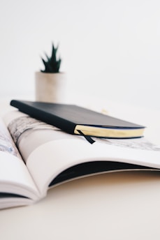 A black notebook is placed on top of an open magazine on a light-colored table. In the background, there is a small plant in a white pot, slightly out of focus.