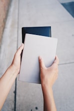 Two hands are holding a grey notebook and a black leather-bound book against a concrete pavement background.