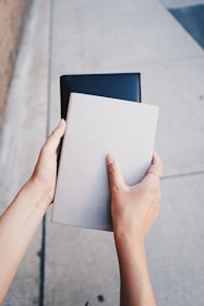Two hands are holding a grey notebook and a black leather-bound book against a concrete pavement background.