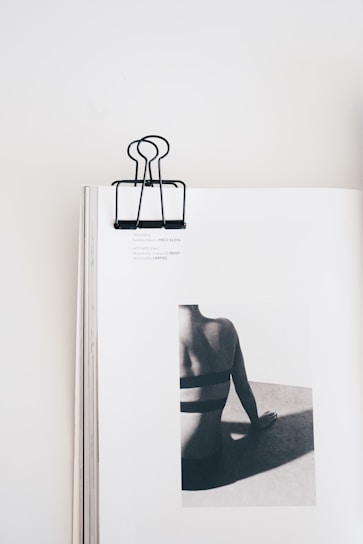 A minimalist composition featuring an open book with a black binder clip on the top center. The book displays a monochrome photograph of a person from the back, sitting and leaning on one hand. The focus is on the person's back and shadows, creating a striking contrast on the page.