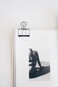 A minimalist composition featuring an open book with a black binder clip on the top center. The book displays a monochrome photograph of a person from the back, sitting and leaning on one hand. The focus is on the person's back and shadows, creating a striking contrast on the page.