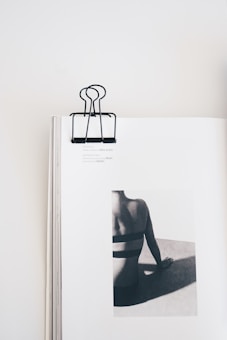 A minimalist composition featuring an open book with a black binder clip on the top center. The book displays a monochrome photograph of a person from the back, sitting and leaning on one hand. The focus is on the person's back and shadows, creating a striking contrast on the page.