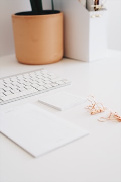 A minimalist desk setup featuring a white keyboard, a large notepad, a smaller sticky note pad, and several rose gold binder clips. A plant in a terracotta pot is partially visible in the background alongside a white organizer holding pens.