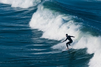 A surfer skillfully rides a large ocean wave, showcasing balance and agility. The wave is breaking with frothy white water, and the surrounding sea is a deep blue.