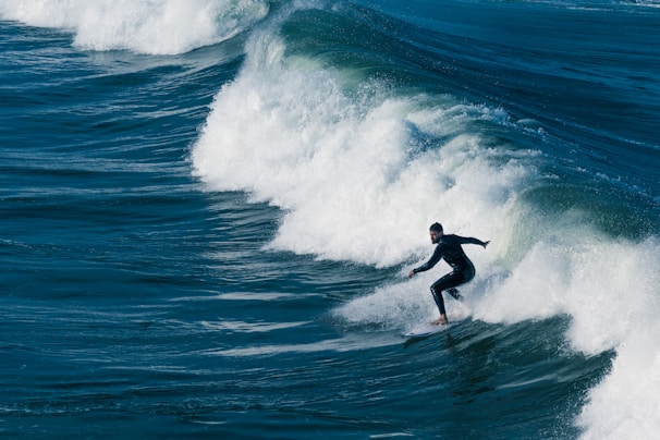 A surfer skillfully rides a large ocean wave, showcasing balance and agility. The wave is breaking with frothy white water, and the surrounding sea is a deep blue.