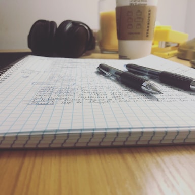 A close-up view of an open notebook with graph paper pages filled with handwritten notes. Two black and silver pens are resting on the notebook. In the background, a pair of headphones, a cup of coffee with a sleeve, and a yellow object are visible. The setting suggests a study or work environment.