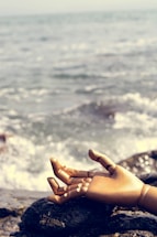 A wooden mannequin hand rests on a rocky surface near the water's edge, with ocean waves gently splashing nearby. The scene blends the organic texture of the rocks with the artificiality of the hand against a serene seascape.