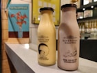 Two bottles of Keventers milkshake are placed on a countertop. The left bottle is a light yellow color with the text 'Thanks for bearing me Mum!' and an image of a bear. The right bottle is a light brown color with the text 'Mum, you're the ducking best!' accompanied by an image of a duck with ducklings. In the background, there are blurred portraits and a bright wall with a blue poster of flowers in a vase.