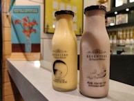 Two bottles of Keventers milkshake are placed on a countertop. The left bottle is a light yellow color with the text 'Thanks for bearing me Mum!' and an image of a bear. The right bottle is a light brown color with the text 'Mum, you're the ducking best!' accompanied by an image of a duck with ducklings. In the background, there are blurred portraits and a bright wall with a blue poster of flowers in a vase.