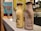 Two bottles of Keventers milkshake are placed on a countertop. The left bottle is a light yellow color with the text 'Thanks for bearing me Mum!' and an image of a bear. The right bottle is a light brown color with the text 'Mum, you're the ducking best!' accompanied by an image of a duck with ducklings. In the background, there are blurred portraits and a bright wall with a blue poster of flowers in a vase.
