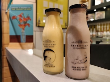 Two bottles of Keventers milkshake are placed on a countertop. The left bottle is a light yellow color with the text 'Thanks for bearing me Mum!' and an image of a bear. The right bottle is a light brown color with the text 'Mum, you're the ducking best!' accompanied by an image of a duck with ducklings. In the background, there are blurred portraits and a bright wall with a blue poster of flowers in a vase.