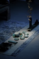 A serene arrangement of traditional tea set pieces is placed elegantly on a patterned rug. The tea set includes various small cups and dishes in shades of green and white. A long, slender vase with dried branches is positioned to the side on a dark tray, contributing to the minimalistic aesthetic. The lighting creates subtle shadows, adding a calm and contemplative atmosphere.