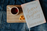 A wooden board rests on a denim fabric surface. On the board, there is a ceramic cup filled with a reddish liquid, likely tea. Next to the cup, a book titled 'Letters to My Daughters: The Art of Being a Wife' by Barbara Rainey is placed. Two small yellow flowers are arranged beside the book.
