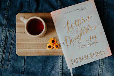 A wooden board rests on a denim fabric surface. On the board, there is a ceramic cup filled with a reddish liquid, likely tea. Next to the cup, a book titled 'Letters to My Daughters: The Art of Being a Wife' by Barbara Rainey is placed. Two small yellow flowers are arranged beside the book.