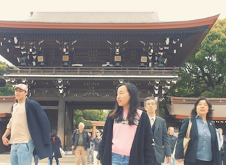 A group of people walk in front of a traditional Japanese gate with a wooden structure. The individuals seem to be casually dressed, with one person in a suit. The background features lush green trees.