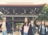 A group of people walk in front of a traditional Japanese gate with a wooden structure. The individuals seem to be casually dressed, with one person in a suit. The background features lush green trees.