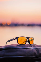 A close-up of a pair of Tapped Sunset sunglasses resting on a sunlit wooden table with a warm orange sunset in the background.