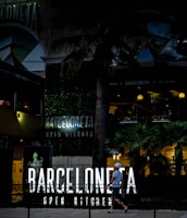 An urban scene featuring a modern restaurant with neon signage reading 'Barceloneta Open Kitchen'. The entrance is surrounded by lush greenery and palm trees. A person walks past the establishment, carrying a drink and wearing casual summer attire, including a hat. The atmosphere is lively, with warm lighting illuminating the outdoor area, suggesting it is an evening setting.