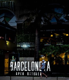 An urban scene featuring a modern restaurant with neon signage reading 'Barceloneta Open Kitchen'. The entrance is surrounded by lush greenery and palm trees. A person walks past the establishment, carrying a drink and wearing casual summer attire, including a hat. The atmosphere is lively, with warm lighting illuminating the outdoor area, suggesting it is an evening setting.