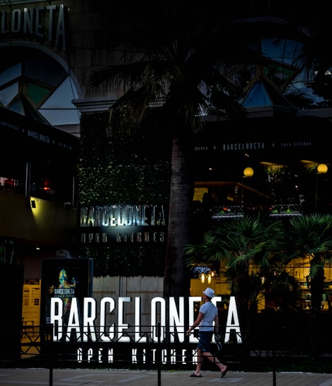 An urban scene featuring a modern restaurant with neon signage reading 'Barceloneta Open Kitchen'. The entrance is surrounded by lush greenery and palm trees. A person walks past the establishment, carrying a drink and wearing casual summer attire, including a hat. The atmosphere is lively, with warm lighting illuminating the outdoor area, suggesting it is an evening setting.