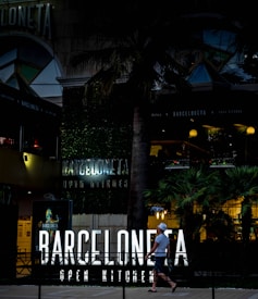An urban scene featuring a modern restaurant with neon signage reading 'Barceloneta Open Kitchen'. The entrance is surrounded by lush greenery and palm trees. A person walks past the establishment, carrying a drink and wearing casual summer attire, including a hat. The atmosphere is lively, with warm lighting illuminating the outdoor area, suggesting it is an evening setting.