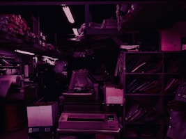 A dimly lit storage room filled with shelves and various office supplies. There are boxes, binders, and paper rolls placed on different racks. A large photocopy machine is visible in the center of the room, surrounded by other electronic equipment. The lighting is primarily fluorescent, casting a purple hue throughout the space.