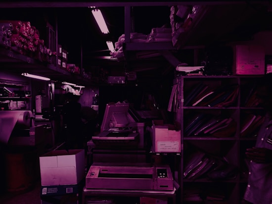 A dimly lit storage room filled with shelves and various office supplies. There are boxes, binders, and paper rolls placed on different racks. A large photocopy machine is visible in the center of the room, surrounded by other electronic equipment. The lighting is primarily fluorescent, casting a purple hue throughout the space.