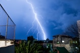 A powerful bolt of lightning strikes against a backdrop of dark blue stormy clouds. The urban setting reveals buildings and a tree, silhouetted against the illuminated night sky. A chain-link fence is visible in the foreground, alongside some foliage.
