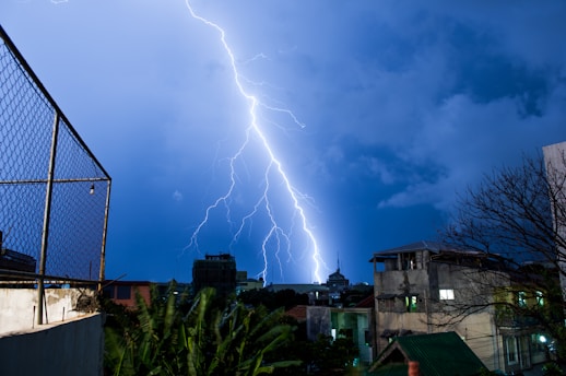 A powerful bolt of lightning strikes against a backdrop of dark blue stormy clouds. The urban setting reveals buildings and a tree, silhouetted against the illuminated night sky. A chain-link fence is visible in the foreground, alongside some foliage.