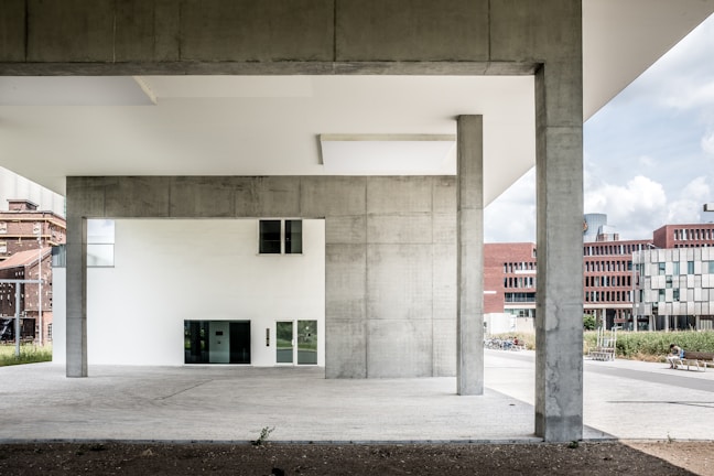 The image features a modern architectural structure with large concrete pillars and minimalistic design elements. In the background, several buildings with red brick and white geometric facades are visible. The foreground is dominated by the open space beneath the structure, which has a clean, urban aesthetic.